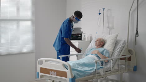 A-black-man-cardiologist-doctor-is-talking-to-an-old-man-patient-lying-on-a-hospital-bed-discussing-rehabilitation-after-recovery.-an-infectious-disease-neurologist-discusses-a-treatment-strategy